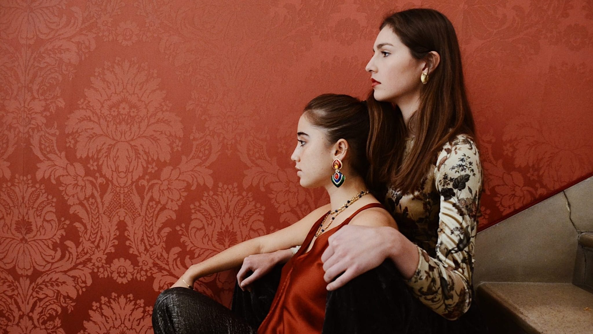 Two girls sit facing away from the camera in front of a red wall