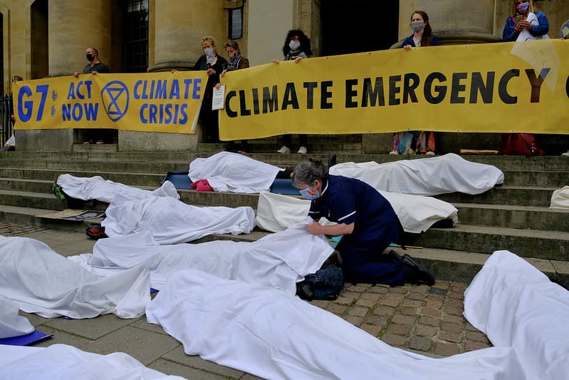 ID: Around seven people lying on the ground completely covered in white sheets. Behind them to the right on steps stand protestors holding yellow banners which read: "G7 / ACT NOW / 'XR LOGO' / CLIMATE CRISIS" and "CLIMATE EMERGENCY". 