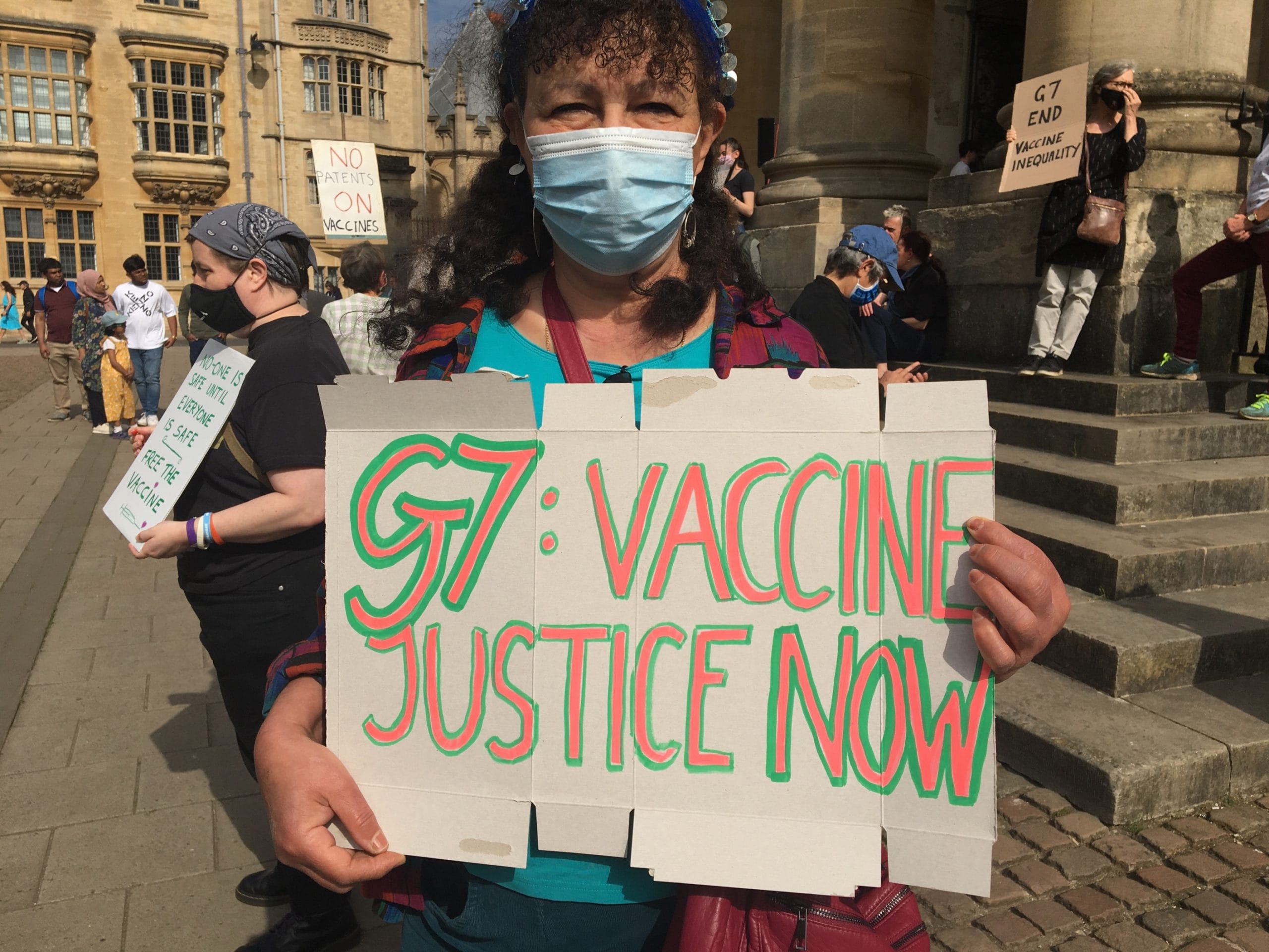 A woman wearing a medical mask holding a sign saying: G7: VACCINE JUSTICE NOW. There are other protestors in the background.